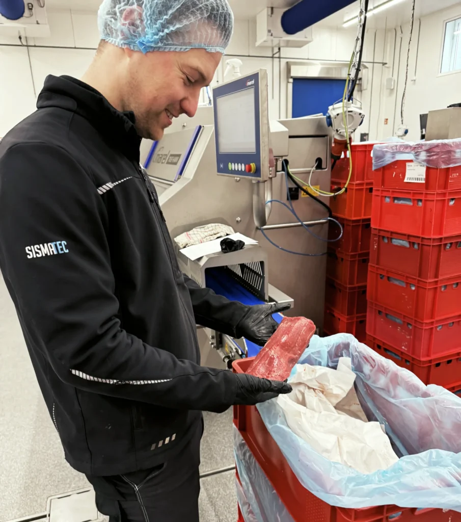 Sismatec specialist inspecting a fresh cut of meat beside a Marel TREIF industrial meat portioning machine.