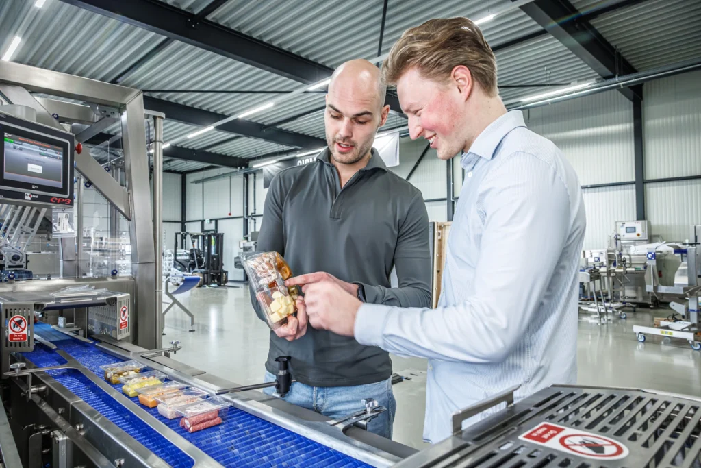 Two Sismatec specialists inspecting sealed convenience food trays beside a Proseal CP3 automatic case packing machine.
