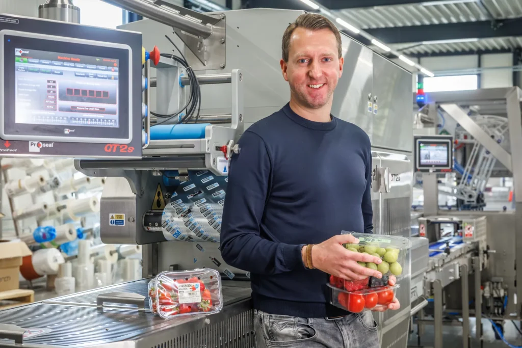 Sismatec specialist holding sealed trays of fresh fruit and vegetables beside a Proseal CT2s automatic tray sealing machine.
