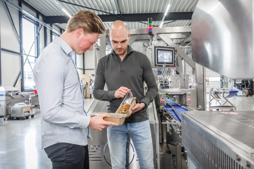 Two Sismatec specialists inspecting sealed food trays beside a Proseal CP3 automatic case packing machine.